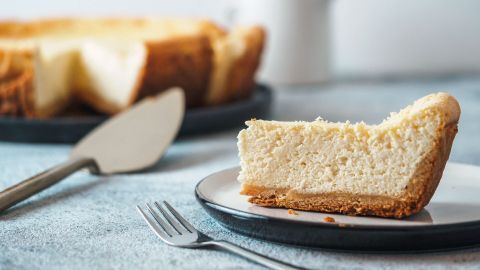 Ein Stück Omas Käsekuchen mit Puddingpulver auf einem Teller, während der gesamte Kuchen im Hintergrund sichtbar ist.