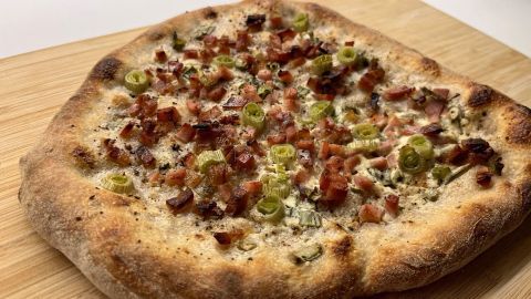 Ein frisch gebackener Flammkuchen ohne Hefe liegt auf einem Holzbrett und ist mit Speck und Schnittlauch belegt.