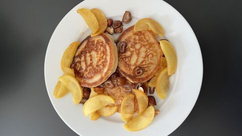 Goldbraune Oladi-Pfannkuchen liegen auf einem weißen Teller, umgeben von frischen Apfelscheiben und Datteln.