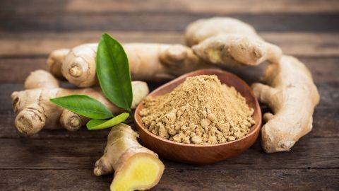 Frische Ingwerwurzeln liegen neben einer Schale voller Ingwerpulver, umgeben von Holztischfläche und grünen Blättern.