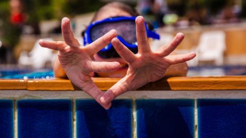 Fingers sind in Wasser sichtbar, während eine Person mit Maske am Rand des Schwimmbeckens spielt und gestikuliert.