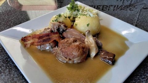 Kalbshaxe liegt saftig in dunkler Biersoße auf einem Teller, umgeben von Kartoffeln und Kraut.