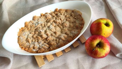 Ein frisch gebackenes Apple Crumble liegt in einer weißen Schale, umringt von Äpfeln auf einem Holzbretthalter.