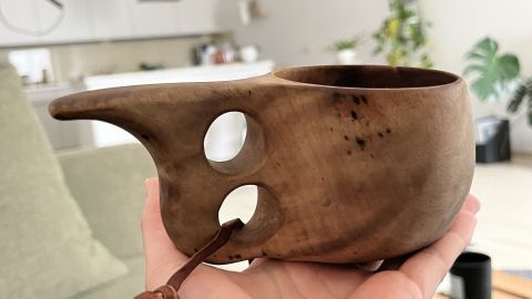 Eine handgefertigte Kuksa wird in einer Hand gehalten, umgeben von einer modernen Wohnküche mit Pflanzen und Dekoration.