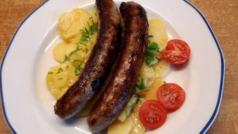 Auf einem Teller liegen zwei gebratene Bratwürste neben Kartoffelsalat und Cherry-Tomaten, angerichtet auf einem Holzuntergrund.