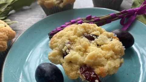 Ein Zwetschgen-Muffin liegt auf einem blauen Teller, umgeben von frischen Zwetschgen und dekorativen Blättern.
