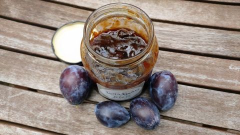 Ein Glas mit selbstgemachtem Zwetschgenmus steht geöffnet auf einem Holztisch, umgeben von frischen Zwetschgen.