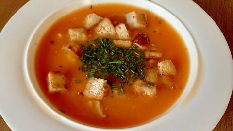 Eine warme Paprika-Kartoffelsuppe ist in einer weißen Schüssel, garniert mit Croutons und frischen Kräutern, auf einem Holzuntergrund.