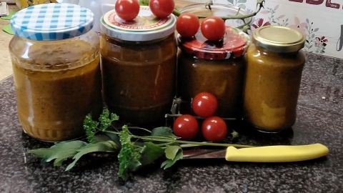 Gläser mit Gemüsepaste stehen neben frischen Tomaten und einem Messer auf einer dunklen Arbeitsplatte in einer Küche.