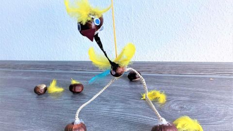 Eine bunte Kastanienmarionette mit Federn steht auf einem Tisch, während einige Kastanien daneben liegen.