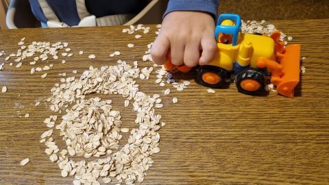 Eine Hand bedient einen kleinen Bagger aus Kunststoff, der Haferflocken auf einem Tisch bewegt, während ein Kind spielt.