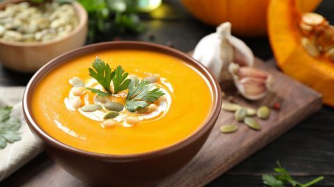 Eine Schüssel mit cremiger Kokos-Kürbissuppe, garniert mit Kürbiskernen und frischem Koriander, steht auf einem Holztisch.