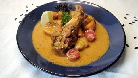 Putenflügel liegen in einer Sauce neben Kartoffeln, Möhren und Kirschtomaten auf einem blauen Teller.