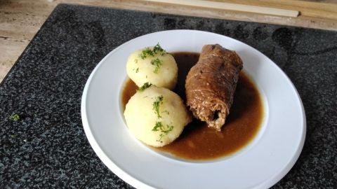 Auf dem Teller liegen zwei Kartoffelknödel neben einer Rinderroulade in brauner Sauce, garniert mit Kräutern.