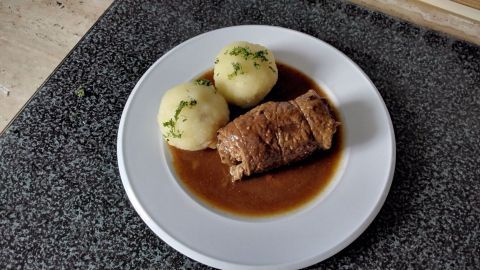 Ein Teller mit Rinderroulade in dunkler Bratensoße, umgeben von zwei Kartoffeln, auf einem grauen Arbeitsfläche.