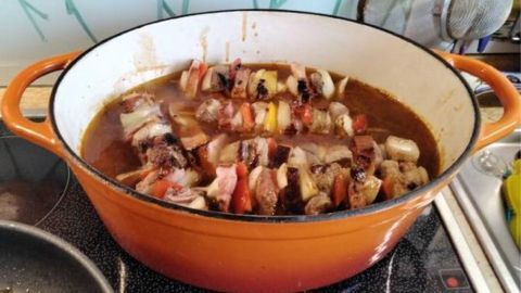 Im großen orangefarbenen Topf simmern Schaschlik mit Gemüse in pikanter Soße auf dem Herd in einer modernen Küche.