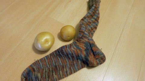 Gegen Halsschmerzen hilft ein DIY-Hausmittel: Kartoffeln kochen, zerdrücken, in einen Wollstrumpf packen und als Wickel um den Hals legen. Die wohltuende Wärme lindert Schmerzen und fördert die Heilung. Ein gestreifter Strumpf liegt auf Holzboden, daneben zwei rohe Kartoffeln.