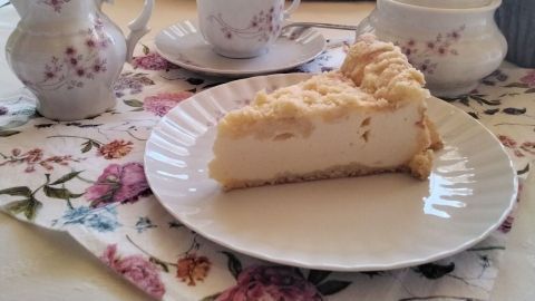 Ein Stück Quarkkuchen mit Streuseln liegt auf einem weißen Teller, umgeben von elegantem Geschirr und einer blumigen Serviette.