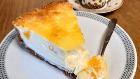 Ein Stück Käse-Schmand-Kuchen mit Keksboden liegt auf einem Teller, während eine Tasse Espresso im Hintergrund steht.