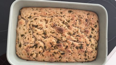 Sauerteig-Focaccia liegt goldbraun in einer quadratischen Auflaufform auf einer Arbeitsfläche und ist mit Kräutern bestreut.