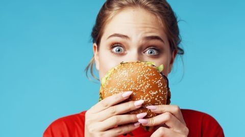 Eine junge Frau hält einen Burger mit Sesambrötchen vor ihrem Gesicht, während sie aufgeregt in die Kamera schaut.