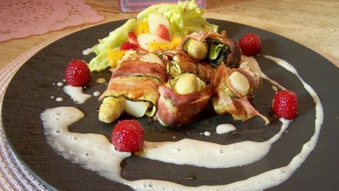 Zarte Spargelröllchen, umwickelt mit Bacon, liegen auf einem schwarzen Teller, umgeben von frischem Salat und Sauce.
