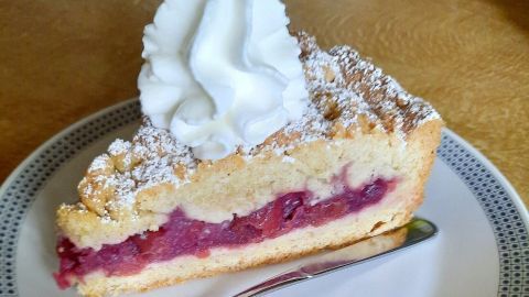 Ein Stück Kirschstreuselkuchen mit Dinkelvollkornmehl und Sahnehaube liegt auf einem Teller, umgeben von Holzoberfläche.