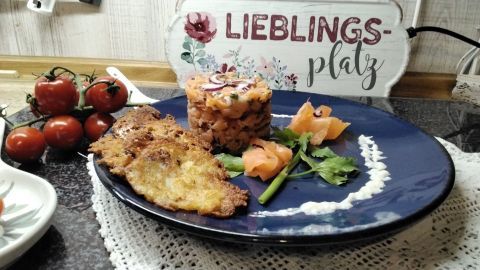 Lachstatar wird ansprechend auf einem blauen Teller mit knusprigen Kartoffelpuffern und frischen Zutaten angerichtet.