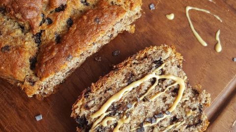 Ein frisch gebackenes, veganes Bananenbrot liegt auf einem Holzbrett, mit einem Stück, das mit einer süßen Soße dekoriert ist.