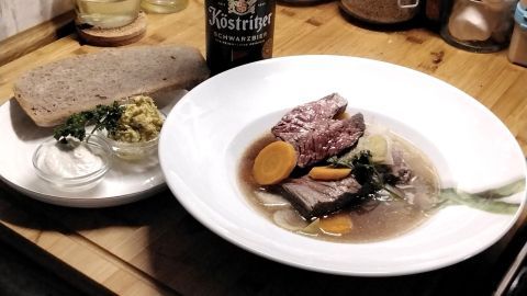 Ein Teller mit Kronfleisch im Sud, umgeben von buntem Gemüse, Brot und Bier auf einer Holzoberfläche.