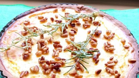 Eine gebackene Orangen-Tarte mit Ricotta und gerösteten Pinienkernen liegt auf einem grünen Tuch, dekoriert mit Kräutern.