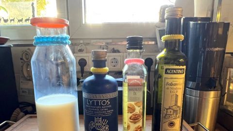 Eine Glasflasche mit Milch und drei Ölfalschen stehen nebeneinander auf einem Tisch mit einem Tuch in einer hellen Küche.