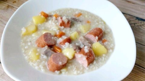 Eine Schüssel Graupensuppe mit Würstchen und Gemüsestücken auf einem Holztisch. Sie wirkt herzhaft und einladend.