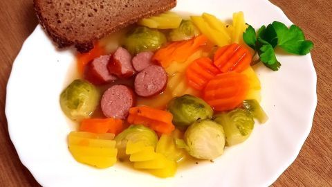 Eine weiße Porzellanteller zeigt eine Gemüsesuppe mit Cabanossi und Gemüse, begleitet von einer Scheibe Brot, auf einem Holzuntergrund.