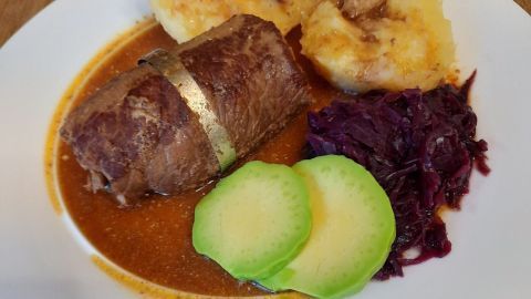 Zarte Rinderroulade liegt in einer Sauce auf einem weißen Teller, umgeben von Kloß, Rotkohl und Gurkenscheiben.