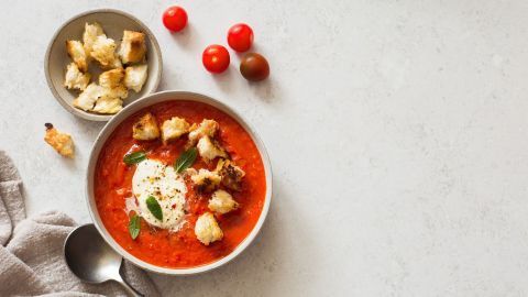 Eine Schüssel Tomatenrahmsuppe mit frischen Kräutern und geröstetem Brot neben einer Schale Kirschtomaten.