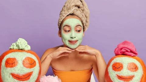 Eine Frau mit einer Gesichtsmaske lächelt charmant zwischen zwei dekorierten Kürbissen, die Fruchtbarkeit im Herbst symbolisieren.