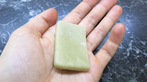 Olivenölseife ist eine weiche und gesunde Alternative zu Shampoos und anderen Seifen für den ganzen Körper. Eine Hand hält ein Stück Olivenseife, das leicht glänzt, auf einer dunklen, marmorierten Oberfläche.