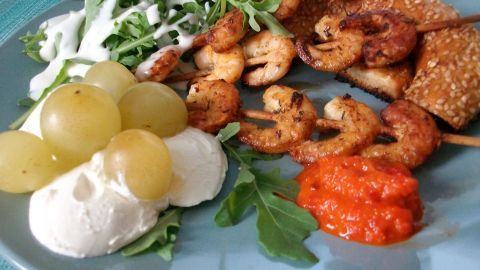 Garnelenspieße liegen auf einem Teller mit Ricotta, Trauben und Rucola, umgeben von frischem Pesto und Dips.