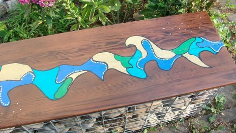 Eine kunstvoll gestaltete Gabionenbank mit einer bunten, wellenförmigen Malerei steht in einem grünen Garten.