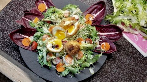 Ein frischer Salat dekoriert mit frittierten Eiern, die mittig auf einer dunklen Platte angeordnet sind, umgeben von Gemüse.