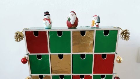 Ein Adventskalender mit Holzschubladen steht dekoriert auf einem Tisch, geschmückt mit Weihnachtsfiguren und Kugeln.