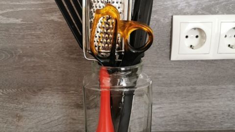 Eine Glasbehälter steht auf einer Arbeitsfläche und hält Küchenutensilien wie einen Rührbesen und eine Reibe.