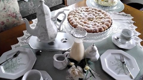 Ein schön gedeckter Tisch zeigt eine große Buchteln-Torte mit Puderzucker, daneben eine Kaffeekanne und Vanillesoße.