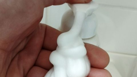 Eine Hand drückt einen Seifenspender und gibt Schaumseife ab, während der Hintergrund eine helle Wand zeigt.