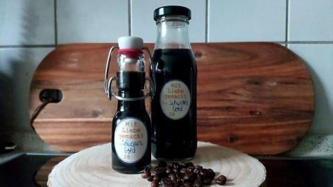 Zwei Flaschen mit Kaffeesirup stehen auf einem Holzbrett, umgeben von Kaffeebohnen in einer gemütlichen Küchenszene.
