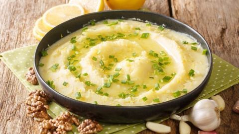 Ein cremiger Knoblauch-Kartoffel-Dip ist in einer schwarzen Schüssel mit grünen Zwiebeln und Zitrone angerichtet, umgeben von Walnüssen.