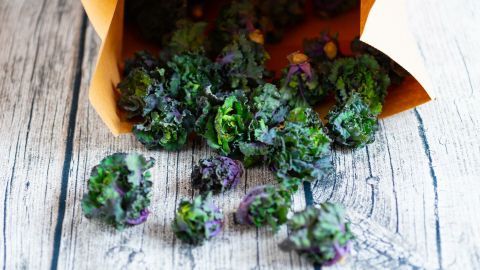 Kale-Blüten fallen aus einer braunen Tüte auf einen rustikalen Holztisch und zeigen ihre grüne und violette Färbung.