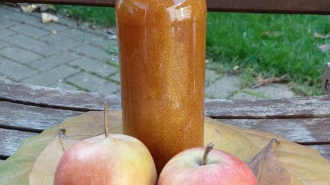 Zwei Äpfel und eine Flasche Bratapfel-Likör stehen auf einem Holztisch, umgeben von herbstlichen Blättern im Garten.