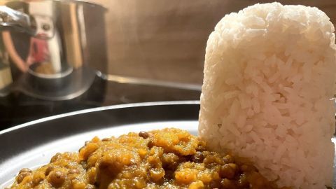 Eine Portion Karotten-Linsen-Curry liegt auf einem Teller neben stabil gedrückt gestapeltem Reis in einer hellen Küche.